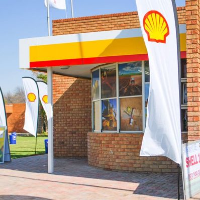 Shell gas station displaying flags and banners, with a feather flying banner attached to a black pole sleeve